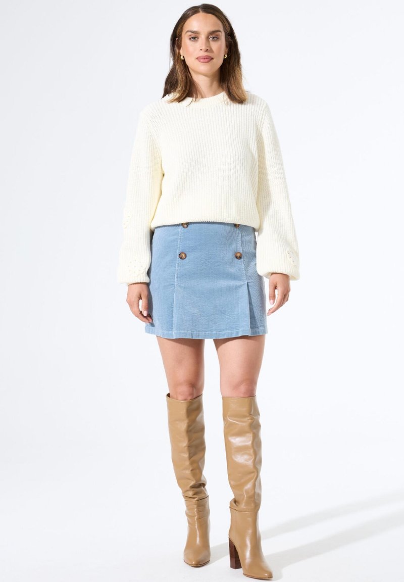 Vrouw in een witte gebreide trui, een blauwe knopen minirok en tan leren laarzen tot aan de knie, staande voor een witte achtergrond.