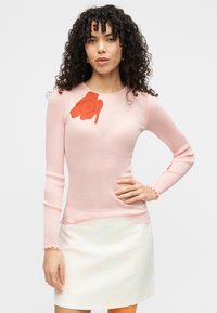 Ροζ ριμπ πουλόβερ με μακριά μανίκια και κόκκινο σχέδιο λουλουδιού στον αριστερό ώμο. Διαθέτει βολάν στο τελείωμα και στις άκρες των μανικιών. Συνδυάζεται με κρεμ φούστα.