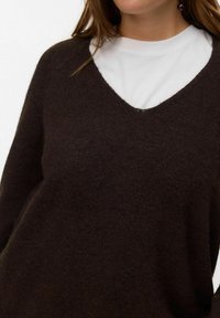 Vrouw met een donkerbruine V-halstrui over een witte ronde hals trui, met lang bruin haar en zichtbare donkere ronde oorbellen.