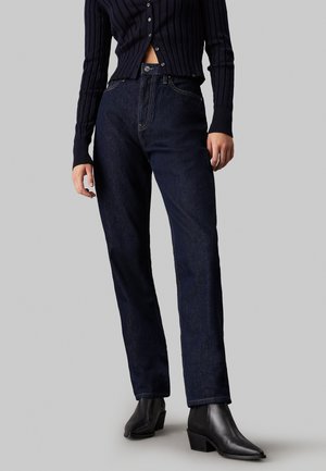 Vaqueros de denim azul oscuro con corte recto, que presentan sutiles detalles de costura y un cierre de botón. Se llevan con un suéter negro de canalé ajustado y botas negras hasta el tobillo.
