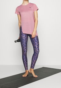 Woman standing barefoot on a yoga mat wearing a pink Puma t-shirt and blue leopard print leggings, holding a black water bottle.