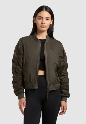 Nő olívazöld bomberdzsekiben, fekete haspólóval és fekete nadrággal, egy egyszínű világos háttér előtt állva.