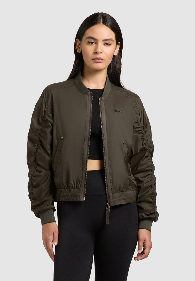 Femme portant une veste bomber vert olive sur un haut court noir et un pantalon noir, debout devant un fond clair uni.