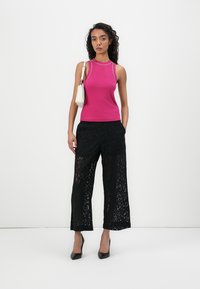 Fuksia tank top tihedalt sobituv mustade pitsiste laiadest pükstest, millel on keerukad mustrid. Mudel hoiab käes väikest valget käekotti.