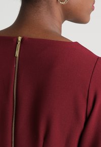 Punaharmaa mekko korkealla kaula-aukolla ja kultaisella vetoketjulla takana. Sileä kangaspinta ja istuva malli.