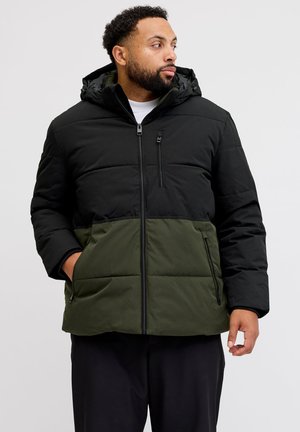 Mann trägt eine schwarze und olivgrüne Steppjacke mit Kapuze, schaut zur Seite, Hände in den Taschen, steht vor einem einfarbigen Hintergrund.