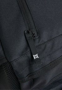 Svart zippered väska med texturerat material och meshdetaljer. Innehåller en liten svart dragkedjehandtag och en etikett med vit text.