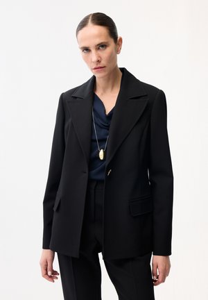 Kvinde med bagstrøget hår iført en skræddersyet sort blazer, marineblå draperet bluse, sorte bukser og et langt guldfarvet vedhængs halskæde.