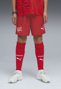 Uniforme atlético rojo con diseño a rayas, con detalles en blanco en los pantalones cortos y calcetas. Incluye la marca Puma y texto en suizo. Los tacos son blancos.