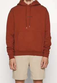 Rostfärgad huvtröja med en känguruficka, justerbara dragsnören och diskret frontlogotyp i svart. Bomullsblandning, avslappnad passform.