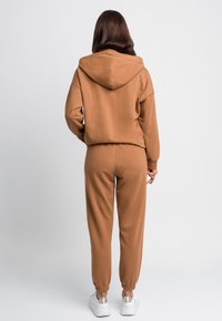 Brązowa bluza z kapturem i dopasowane dresowe spodnie wykonane z miękkiego materiału, z kapturem i elastycznymi mankietami, w połączeniu z białymi trampkami.