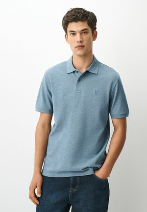 Ung mand stående med den ene hånd i lommen iført en lyseblå kortærmet polo og mørkeblå jeans på en ensfarvet baggrund.