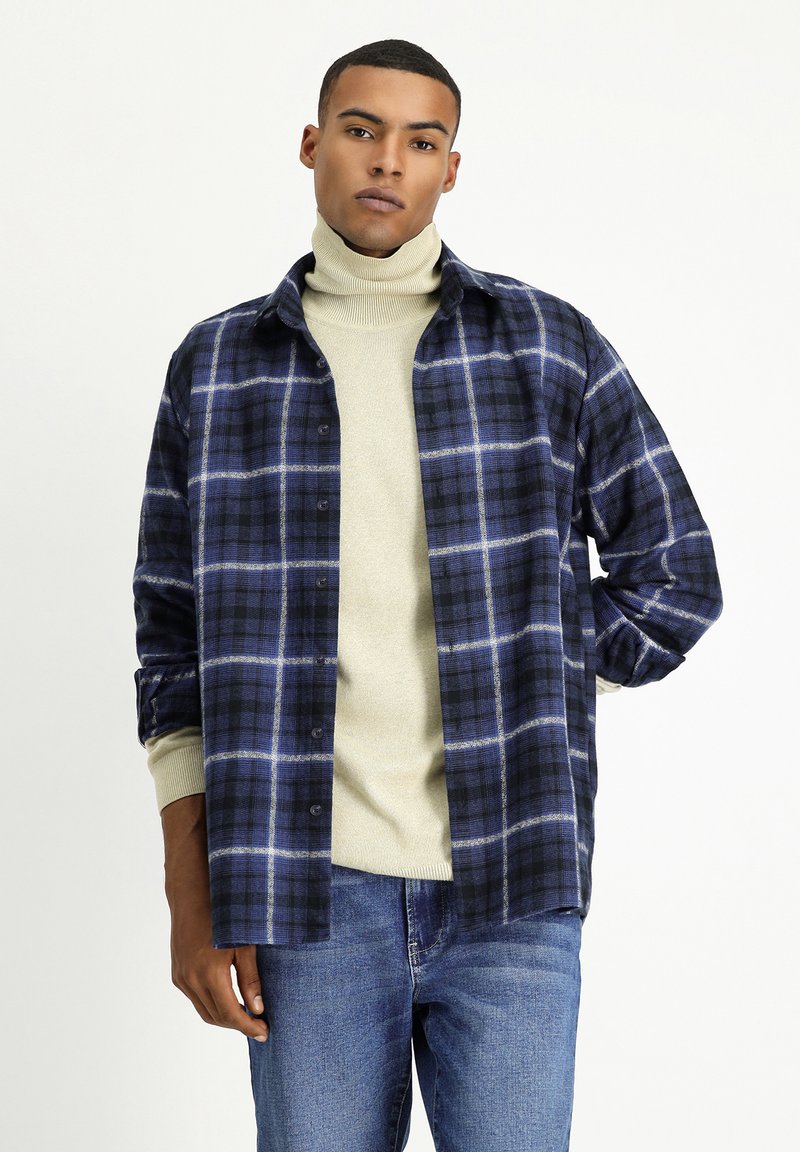 Camicia di flanella a quadri in blu navy e nero con accenti bianchi, con bottoni frontali, maniche lunghe, indossata sopra un pullover a collo alto color beige chiaro.