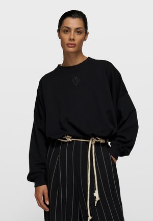 Czarna, oversizeowa bluza z prążkowanym dekoltem i mankietami; z subtelnym haftem na piersi; noszona z ciemnymi spodniami w paski.