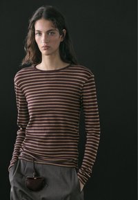 Femme aux cheveux longs et ondulés portant un chemisier à manches longues rayé marron et beige, et un pantalon gris avec une petite pochette marron accrochée à la taille.