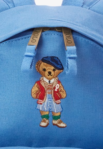 Μπλε σακίδιο πλάτης με κεντημένο αρκούδο που φοράει κόκκινο μπλούζα τύπου varsity, ριγέ μπλούζα, φούστα καρό και πράσινες κάλτσες, με χρυσές φερμουάρ.