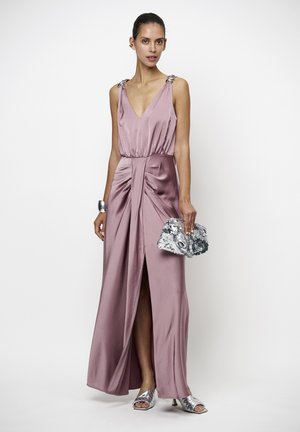 Vrouw die een mouwloze roze satijnen jurk met een voorste split draagt, zilveren open-teen hakken, een zilveren manchettenarmband, en een zilveren pailletten clutch vasthoudt.