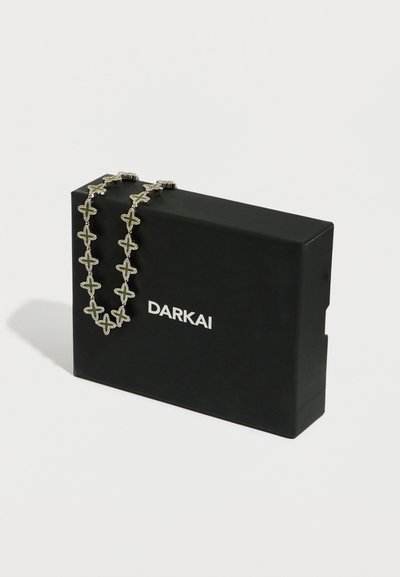 Srebrna ogrlica s povezavami v obliki križa in zelenimi poudarki, prikazana na črni škatli z belim tiskom imena znamke "DARKAI".