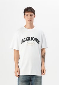 Ung mand med kort, mørkt hår iført en hvid oversized t-shirt med "JACK & JONES" tekst og blå jeans, stående foran en hvid baggrund.