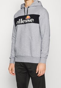 Sweat à capuche gris avec panneau avant noir arborant le logo "ellesse". Capuche ajustable avec cordon de serrage et poche kangourou. Poignets et ourlet côtelés.