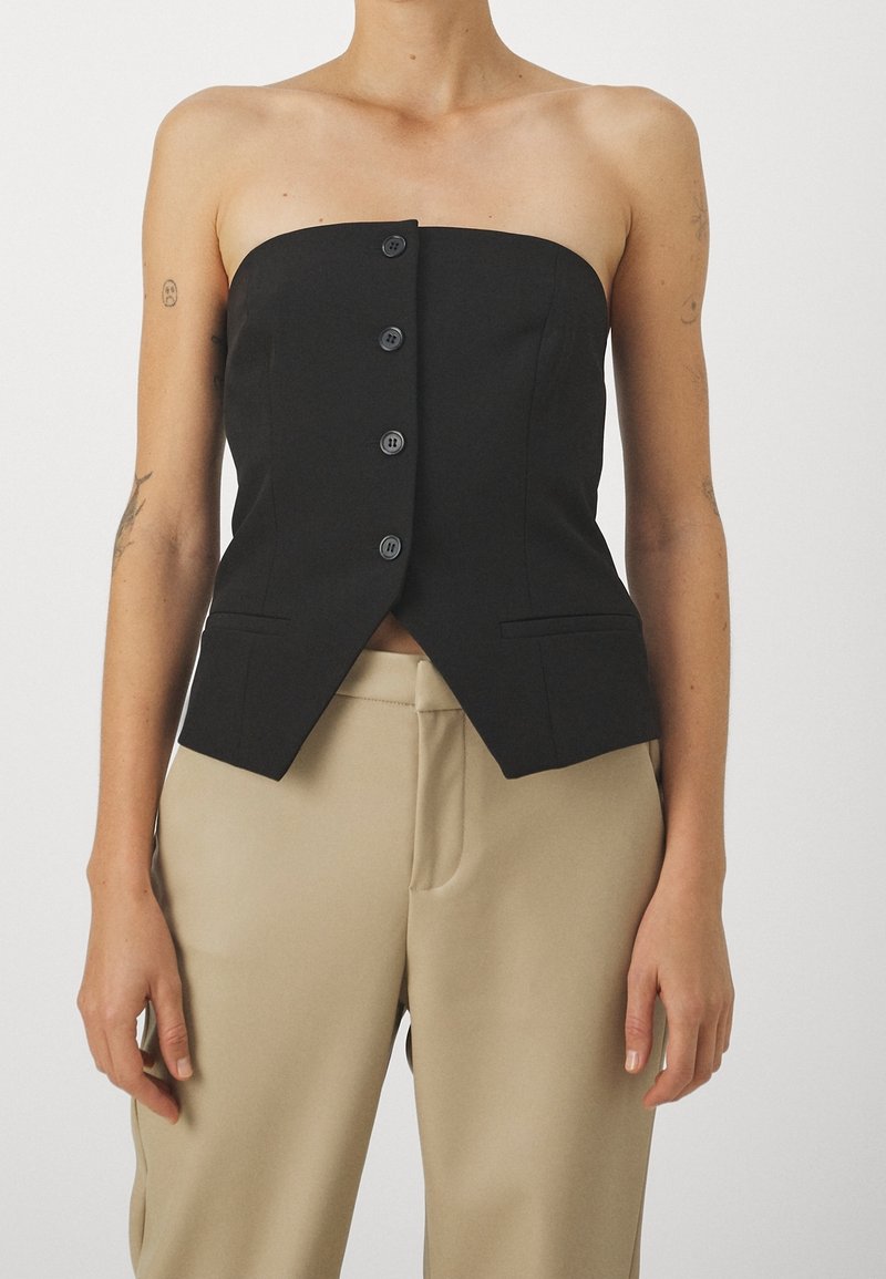 Haut noir sans bretelles avec boutons et ourlet avant pointu porté avec un pantalon beige, bras détendus le long du corps sur fond blanc.