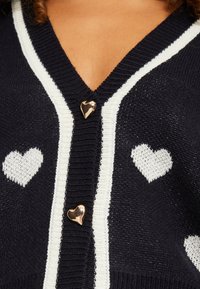 Marineblauwe cardigan met witte hartpatronen; heeft een V-hals en gouden hartvormige knopen aan de voorkant. Zachte brei-textuur.