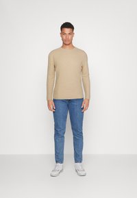 Beige struktureret striktrøje med rund halsudskæring, parret med blå denim jeans og hvide sneakers i en frontvendt position.