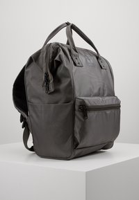 Šedý batoh vyrobený z odolné tkaniny, který má dvojité horní rukojeti, přední kapsu na zip a strukturovaný povrch. Součástí jsou také nastavitelné ramenní popruhy.