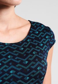 Bluzka w wzory z niebieskim i turkusowym geometrycznym designem na ciemnym tle, z okrągłym dekoltem i krótkimi rękawami.