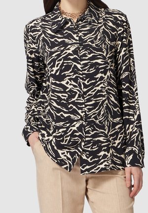Vrouw draagt een zwart-beige blouse met tijgerprint, beige broek en een gelaagde gouden ketting, met één hand in de zak.