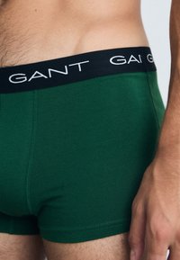 Bóxer verde hecho de una tela suave, con una cinturilla elástica negra que tiene letras blancas. El diseño enfatiza un corte ajustado.