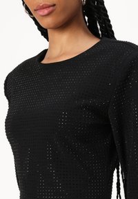 Melnā blūze ar teksturētu virsmu, kurā vienmērīgi izkaisīti mazi, spīdīgi punkti, ar apaļu apkakli un īsām piedurknēm.