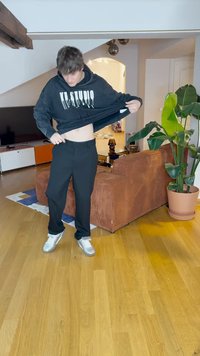 Black hoodie with white dripping text, black pants, and silver sneakers. Indoor setting with wood flooring, a brown couch, and a potted plant.