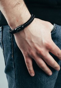 Juodas pintas apyrankė, pagaminta iš patvarios medžiagos, su sklandžiu cilindriniu užsegimu ir išgraviruotu prekių ženklu.