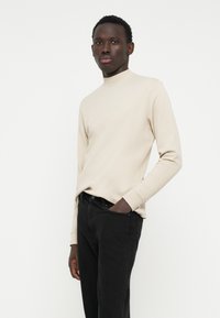 Top beige a maniche lunghe con collo alto, abbinato a pantaloni neri. Il tessuto appare liscio e aderente, con polsini a coste. Design semplice.