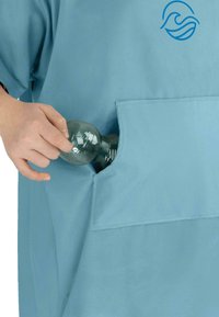 Helles blaues Stoffkleidungsstück mit einer Vordertasche; eine Hand legt einen durchsichtigen grauen Gegenstand in die Tasche. Der Stoff wirkt glatt.