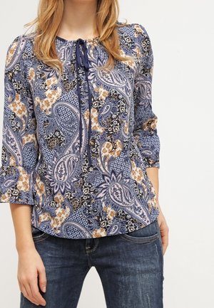 Femme portant un chemisier à motif cachemire bleu, noir et beige avec des manches trois-quarts et un jean bleu foncé.