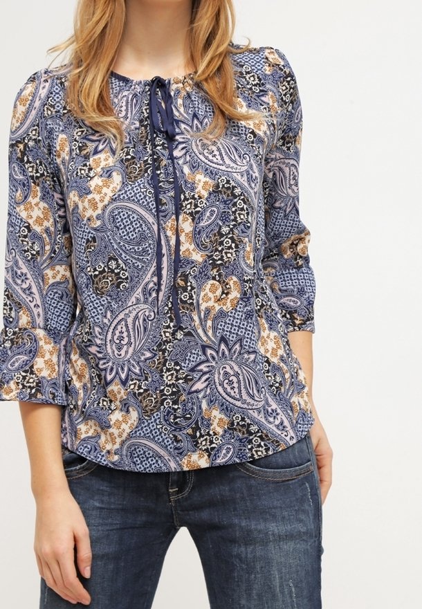 Femme portant un chemisier à motif cachemire bleu, noir et beige avec des manches trois-quarts et un jean bleu foncé.