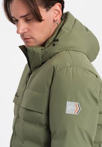 Olivově zelená prošívaná bunda s kapucí, obsahující náprsní kapsu a oranžovo-šedé logo značky na rukávu. Hladký, polstrovaný materiál.
