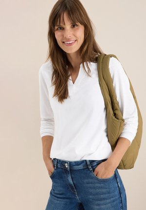Witte lange mouwen top met een V-hals, gecombineerd met een blauwe spijkerbroek. Een lichtgewicht olijfgroene vest hangt over één schouder.