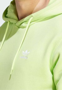 Ljusgrön hoodie med snörning, tillverkad av ett mjukt material, med en vit broderad Adidas-logotyp på bröstet.