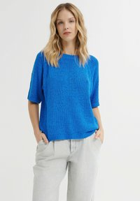 Helles blauer Strickpullover mit kurzen Ärmeln, strukturierter Oberfläche und rundem Halsausschnitt. Kombiniert mit hellgrauen, locker sitzenden Hosen.