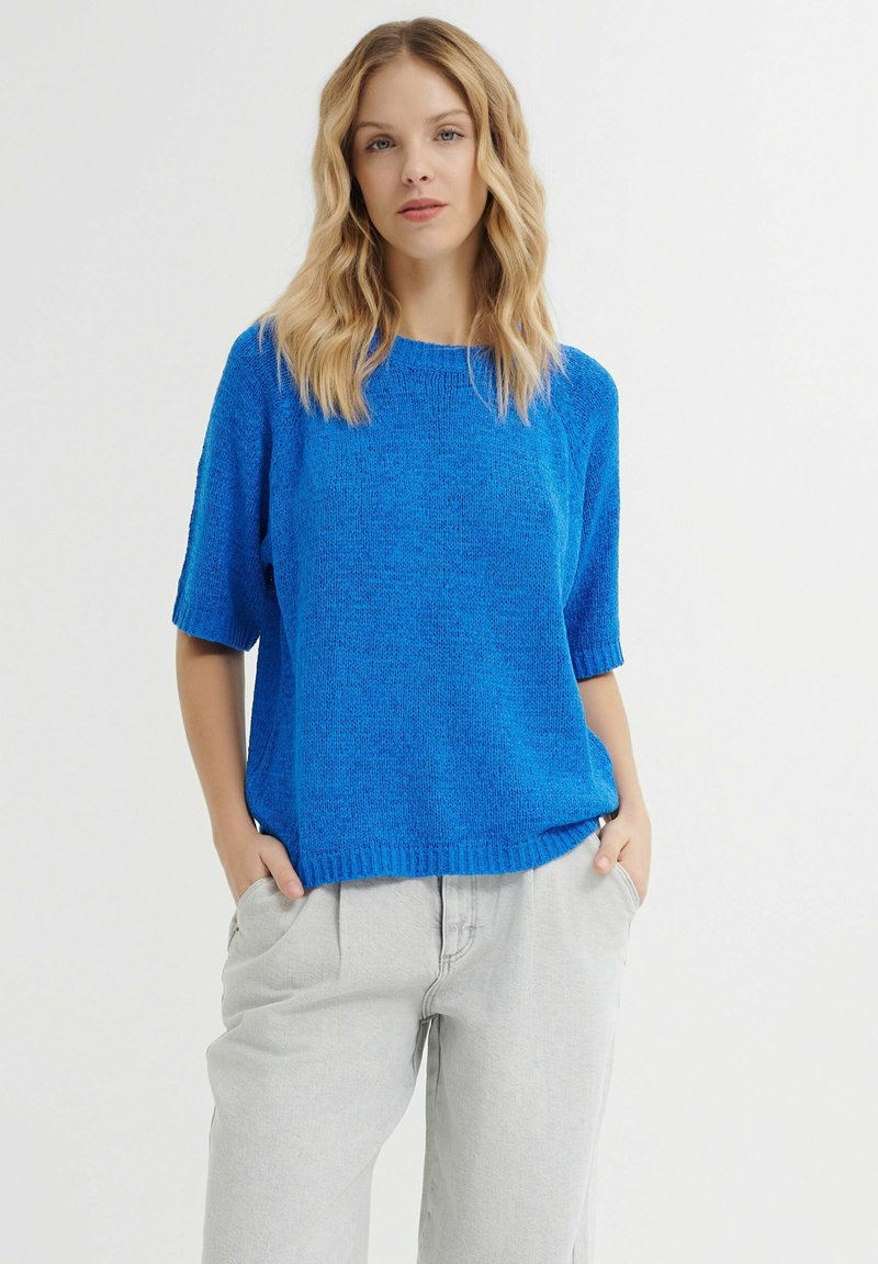 Helles blauer Strickpullover mit kurzen Ärmeln, strukturierter Oberfläche und rundem Halsausschnitt. Kombiniert mit hellgrauen, locker sitzenden Hosen.