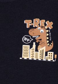 Maglietta navy con un cartone animato di T-Rex in città, con accenti gialli e marroni, una nuvoletta che dice "ROAR" e grafiche di edifici sottostanti.