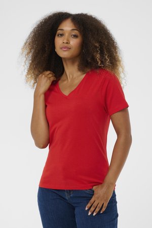 Femme aux cheveux bouclés portant un T-shirt rouge à col en V et un jean bleu, posant avec une main près du cou et l'autre dans la poche.