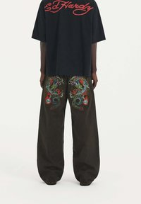 Camiseta negra de corte oversized con el logo rojo de "Ed Hardy", combinada con pantalones holgados oscuros que presentan bordados coloridos de dragones en la parte trasera.