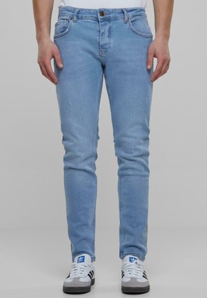 Jeans azul claro de corte slim, con detalles desgastados, cierre de cremallera y bolsillos frontales, combinados con zapatillas grises y blancas.