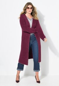 Bordový dlouhý kardigan s otevřeným předním dílem, pletená textura a kontrastní stehy. Nosený přes šedý top a spárovaný s modrými zkrácenými džíny.