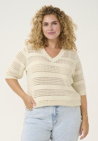 Pull crème en tricot avec un col en V, des manches courtes et un design ajouré présentant des motifs rayés et des accents texturés. Associé à un denim clair.