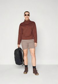 Brun, tekstureret sweatshirt med halv lynlås, beige shorts, sort rygsæk og brune vandrestøvler. Modellen bærer solbriller og står foran en lys baggrund.